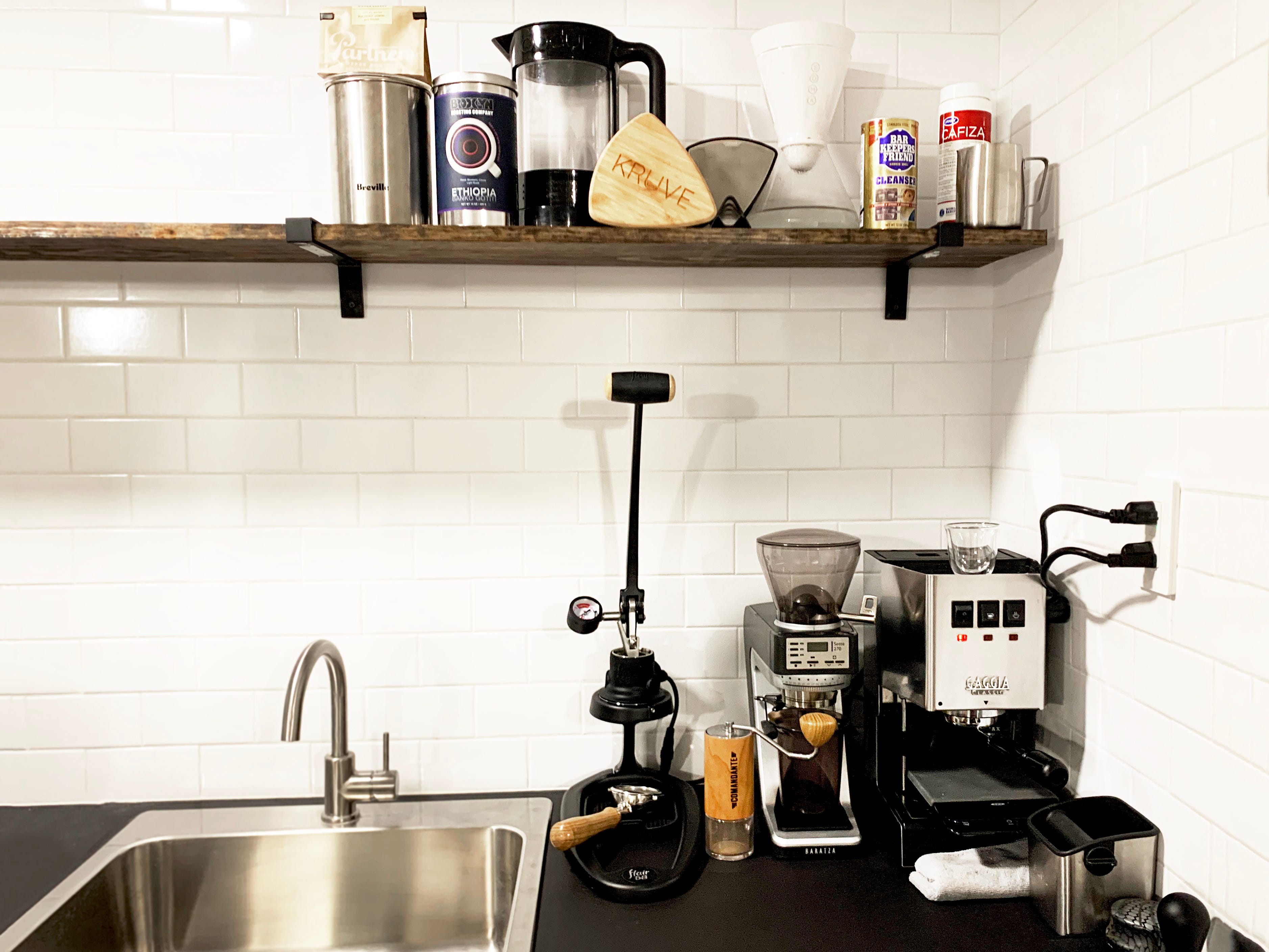 A home espresso bar with multiple machines and grinders on a black countertop in front of white subway tile